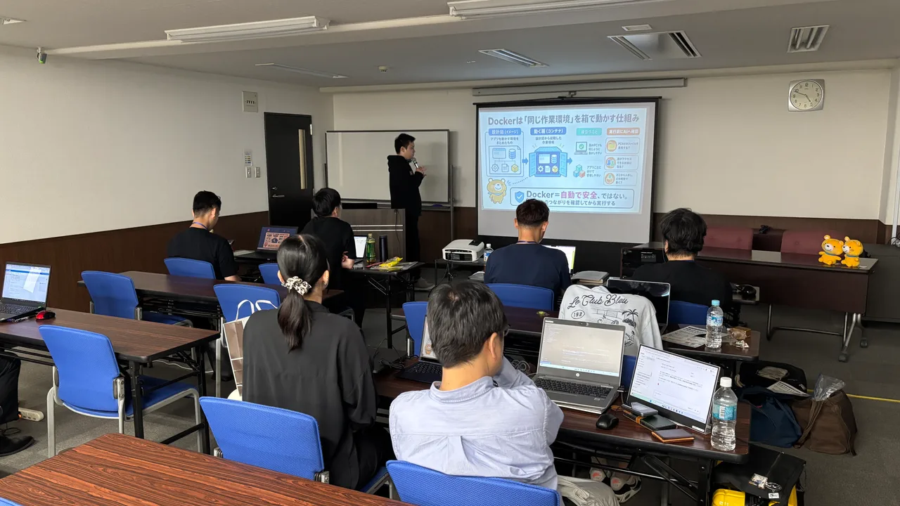 1日目、作業環境を分ける仕組みをスクリーンで説明する中島と、パソコンを開いて受講する参加者