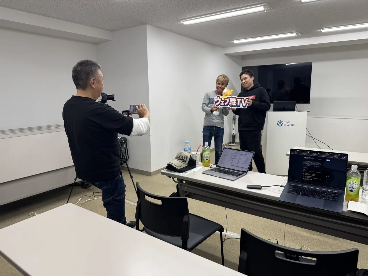 「ウェブ職TV」のパネルを持って中島大介と並ぶ参加者を、別の参加者がスマートフォンで撮影している様子
